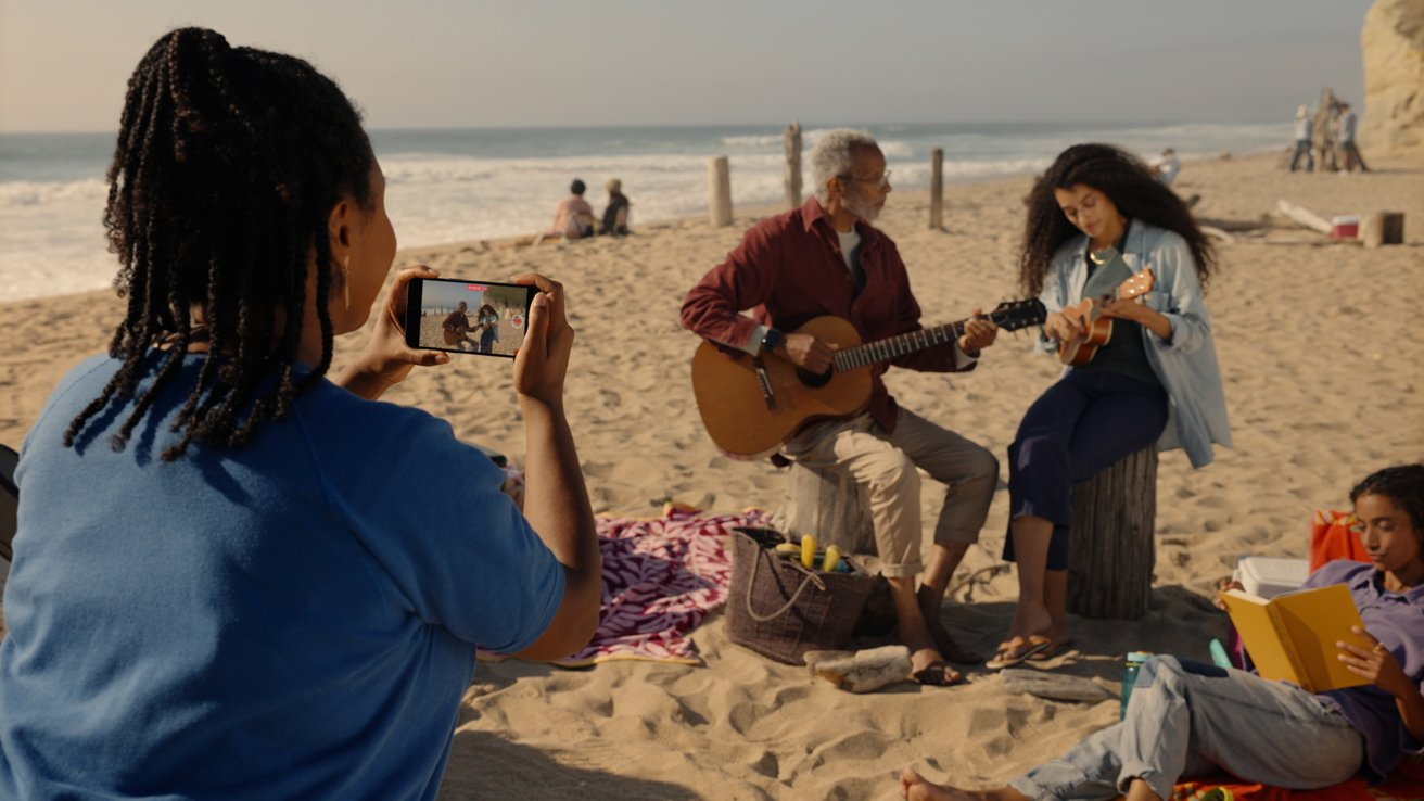 A woman recording a video with her iPhone of two people playing music instruments on a beach.