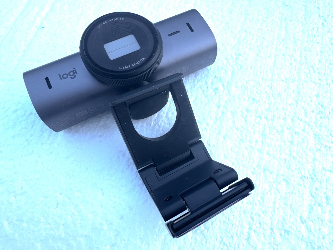 Logitech 4K webcam with lens cap on, mounted on a black monitor clip against a textured light blue background.