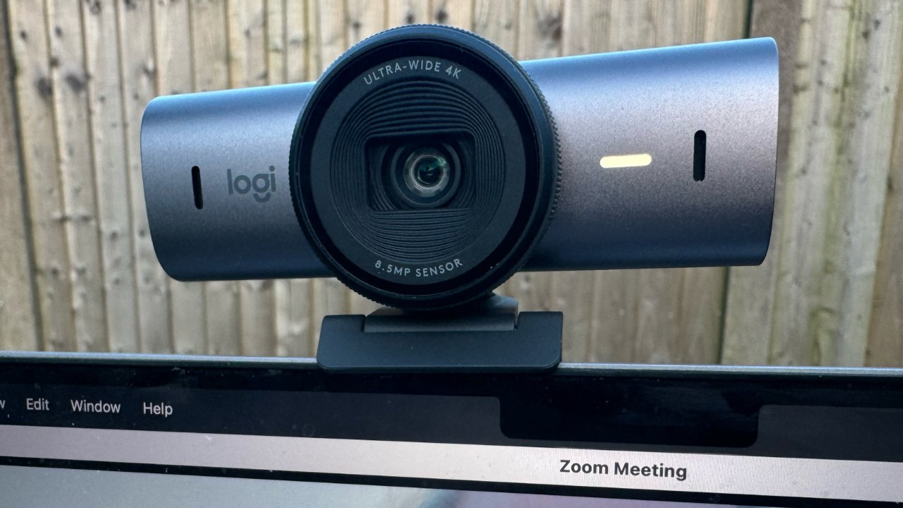 A webcam mounted on a computer monitor displaying the text 'Zoom Meeting' with a blurred wooden fence background.