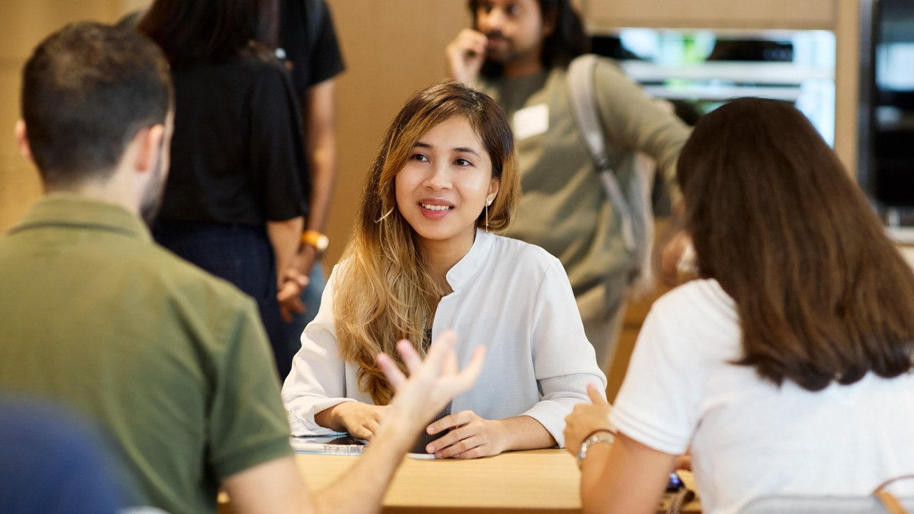 A smiling young woman engaging in a conversation with others around a table in a casual meeting or workshop setting.