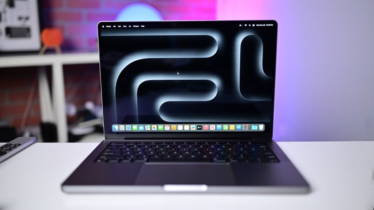 14-inch MacBook Pro with M3 on a desk with colorful blurred background, displaying a dark abstract wallpaper and a dock of application icons.