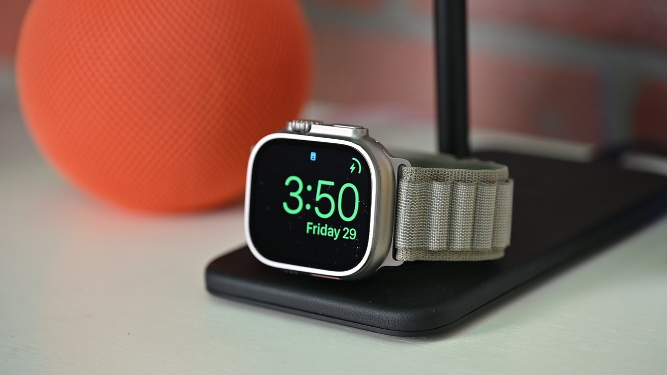Apple Watch Ultra 2 displaying 3:50pm Friday 29 on its face, mounted on a charging stand, with a blurred orange speaker in the background.