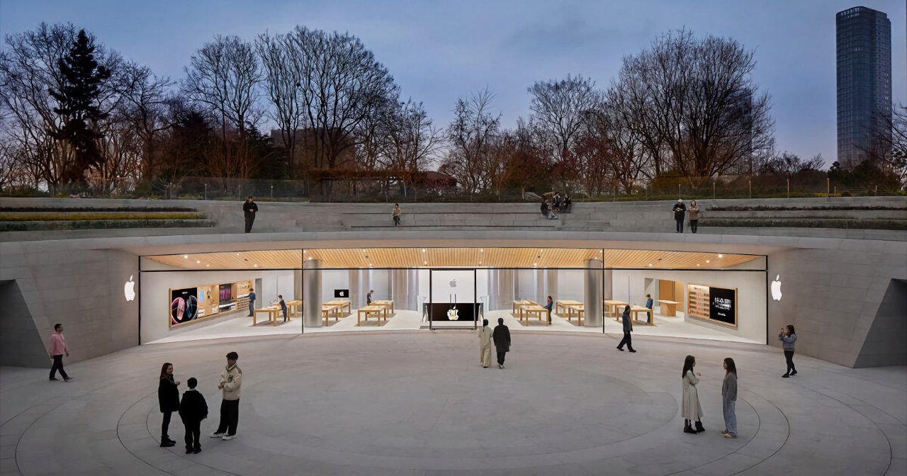 Exterior view of a modern storefront with a lit Apple logo, surrounded by trees and several people milling about at dusk.