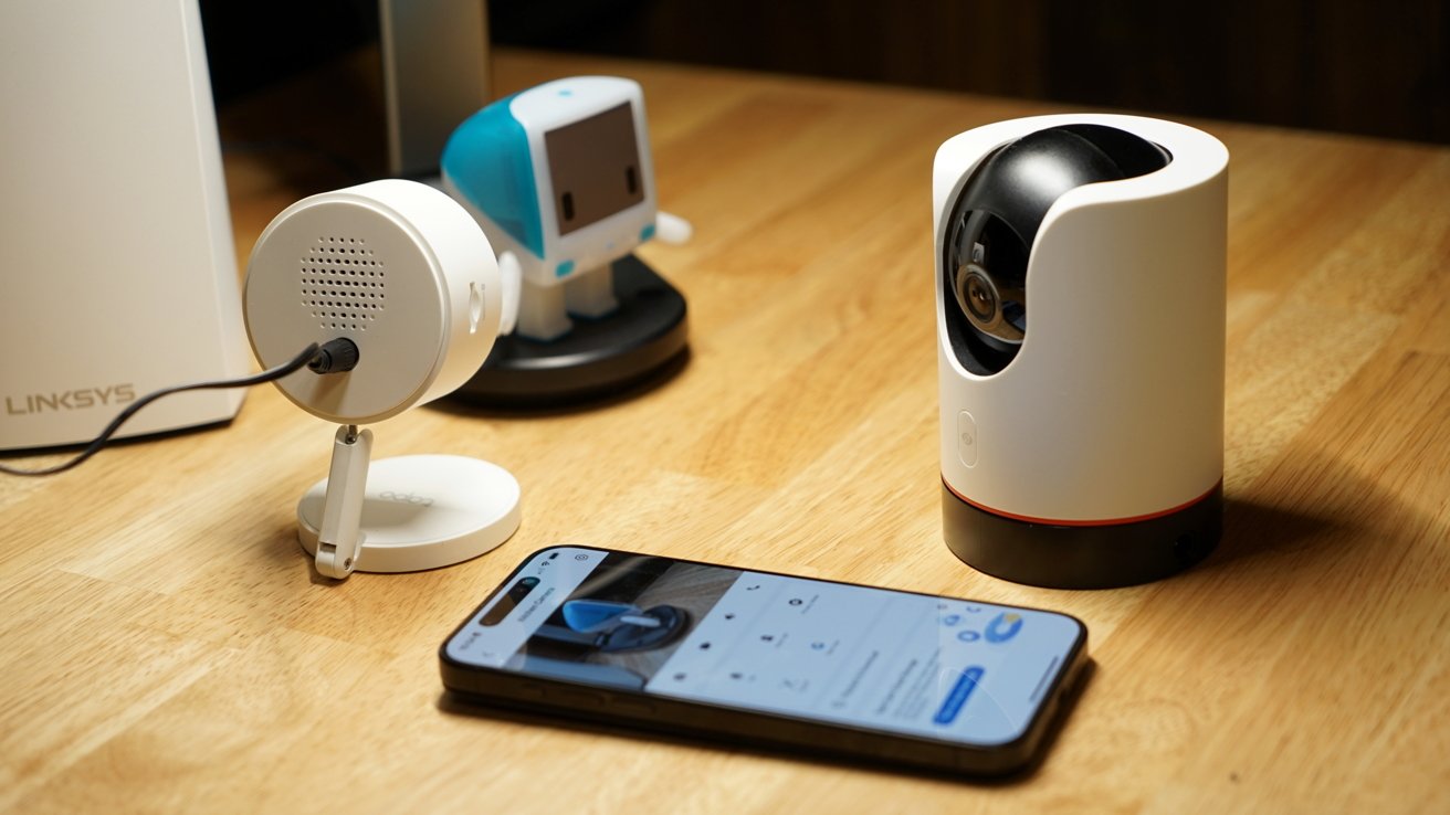 Two home security cameras on a desk. One looks at a small robot toy which is also visible on an iPhone display on the desk.