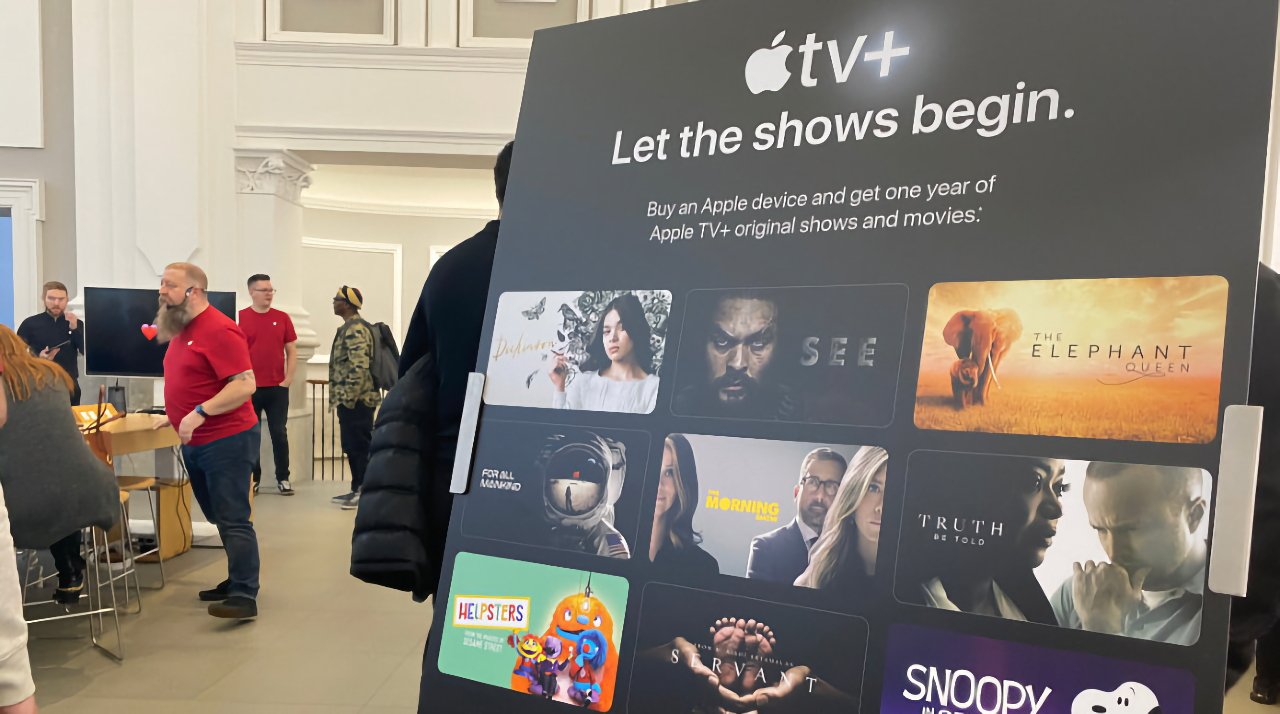 Apple TV+ promotional stand with text and thumbnails for shows, set in a bright store with employees and customers in background.
