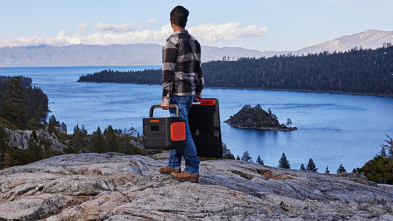 Man standing on rocky overlook with lake view, holding a Jackery portable power station, with pine trees and mountains in the distance.