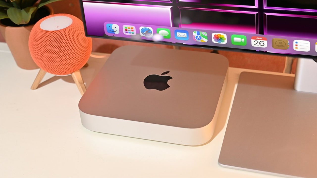 An Apple Mac mini on a desk next to an orange HomePod mini, beneath a monitor displaying a colorful screen saver.