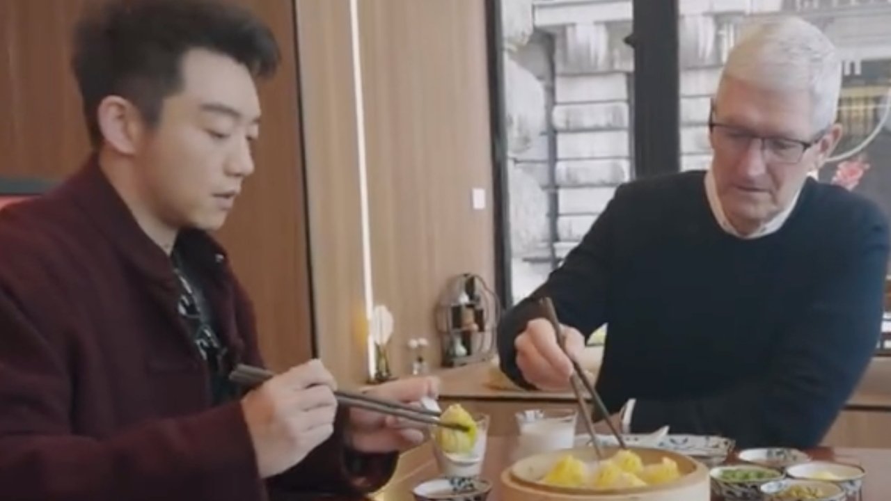 Two men dining, one using chopsticks and the other a fork, with a plate of dumplings between them.