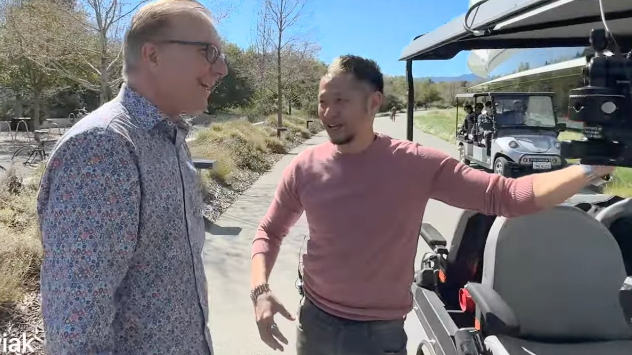 Two men talking beside a golf cart, with a camera mounted on it, outdoors on a sunny day.