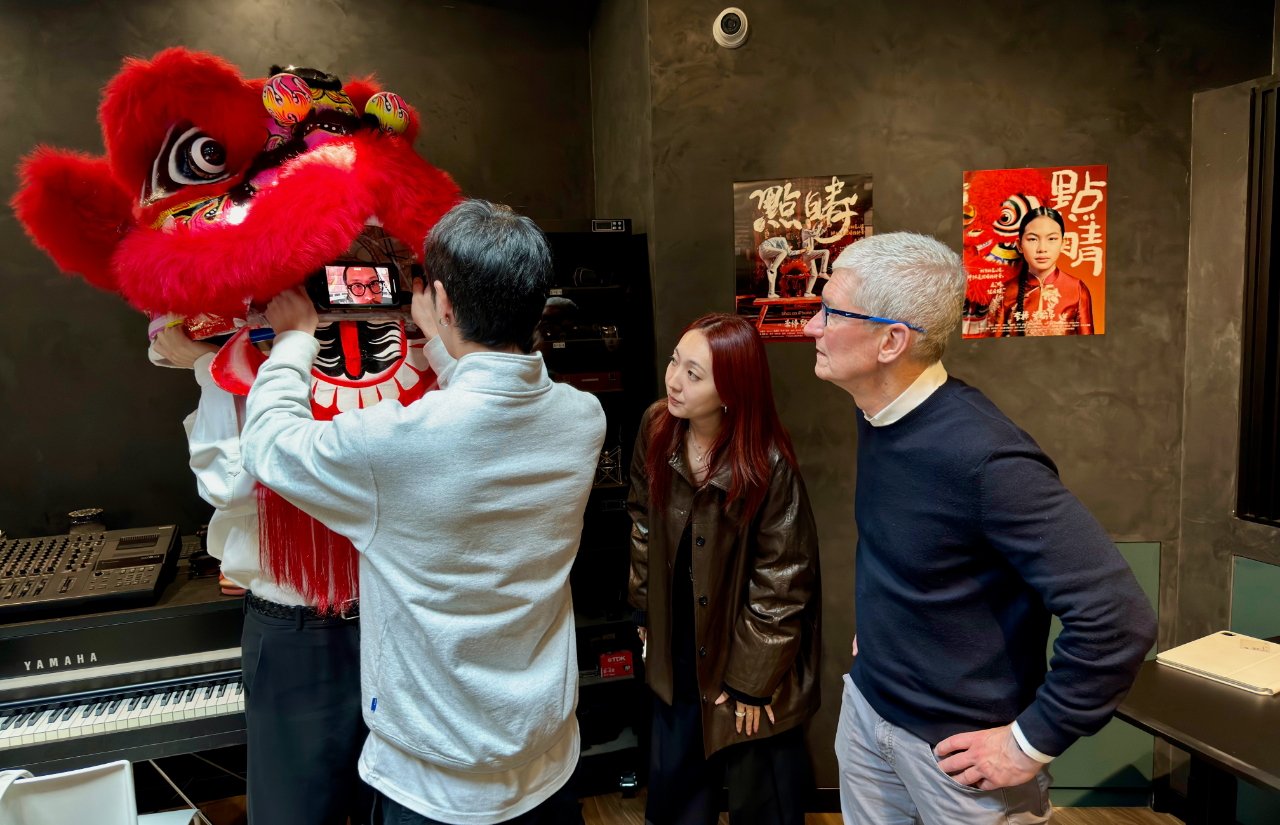 Three people indoors with a red lion dance costume, one holding a phone showing a man's face, musical instruments nearby, and cultural posters on the wall.