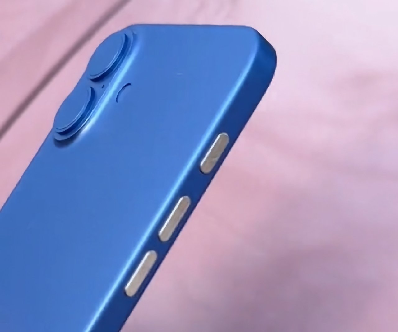 Close-up of a blue smartphone with dual cameras and side buttons, resting on a pink surface.