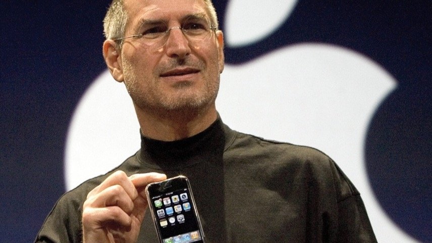 Steve Jobs in black turtleneck presenting a smartphone with a backdrop displaying a partial logo resembling an apple.