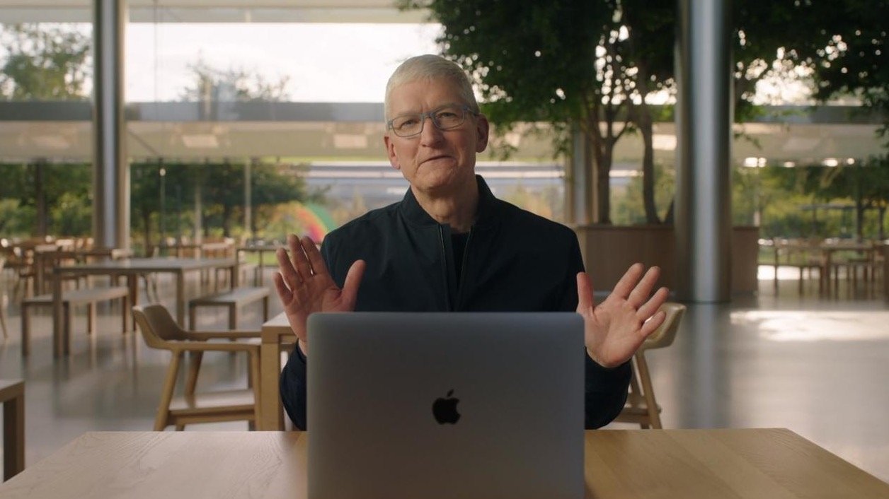 Tim Cook in black shirt sitting at a table with a laptop, gesturing with his hands, in a room with large windows and tables.