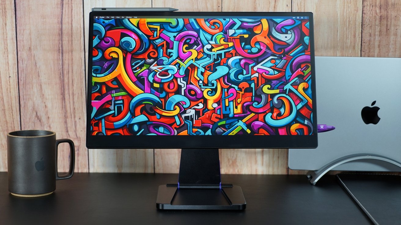 A 17-inch monitor with a colorful wallpaper on a desk next to a MacBook Pro and a mug