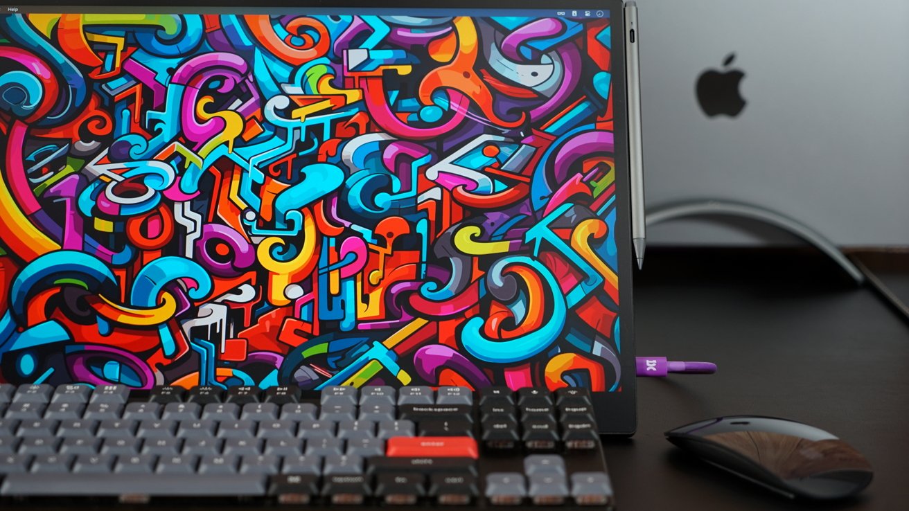 A monitor on a desk with a colorful wallpaper. A keyboard and mouse are in front, and a MacBook Is visible in the background.