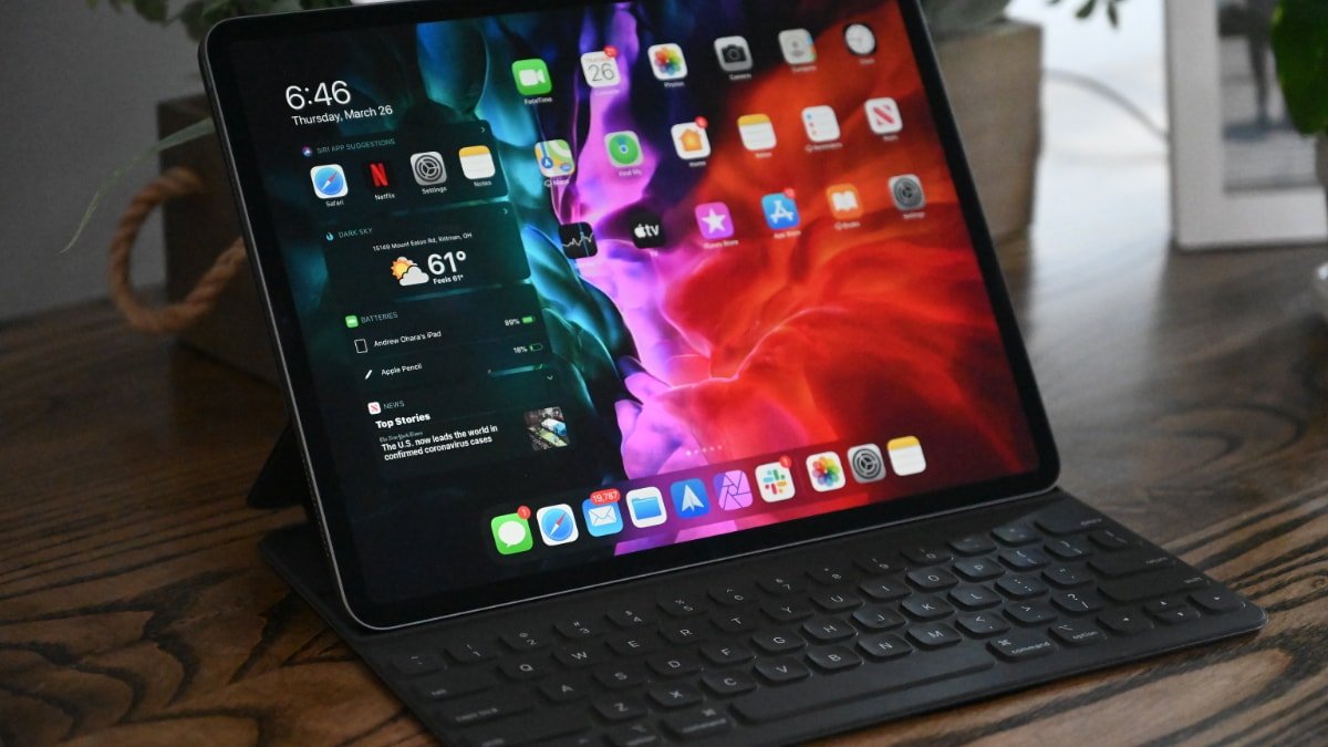 An iPad Pro 2020 with colorful display attached to a keyboard on a wooden surface, displaying various app icons and weather widget.