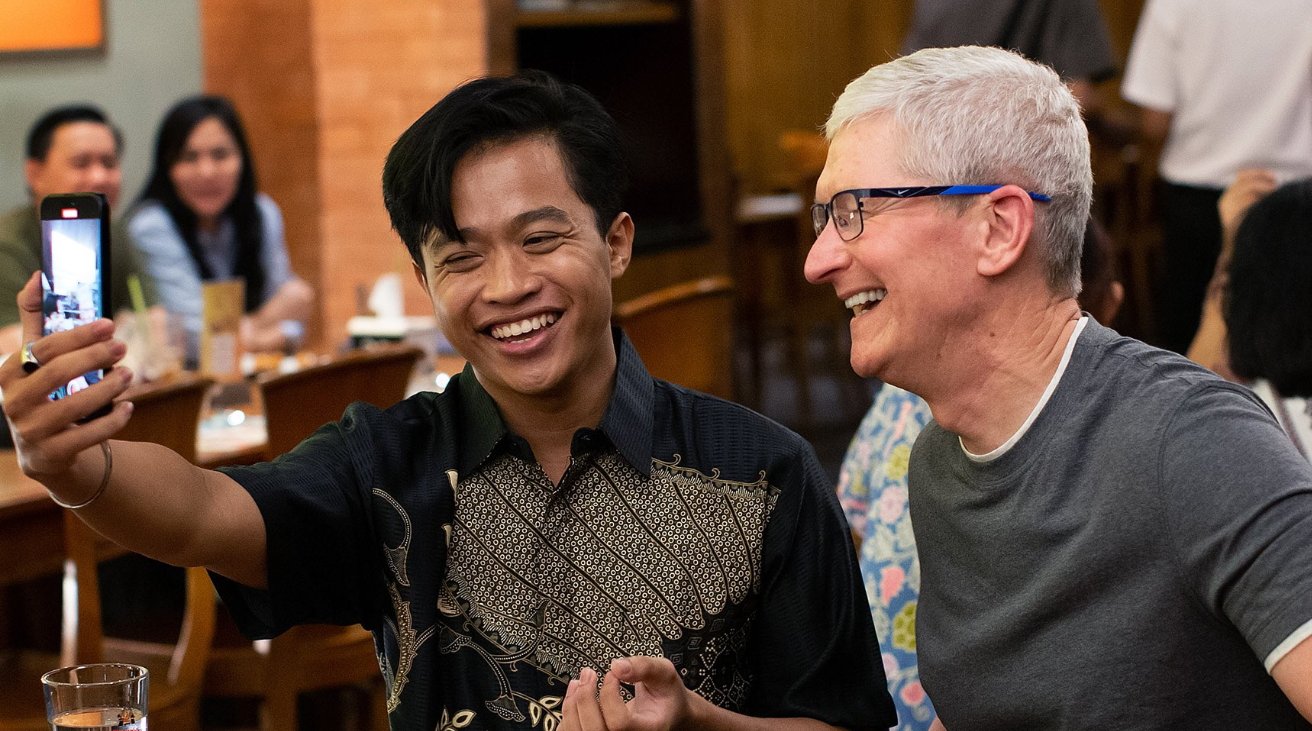 Two men laughing joyfully as one takes a selfie, with others in the background at a dining area.