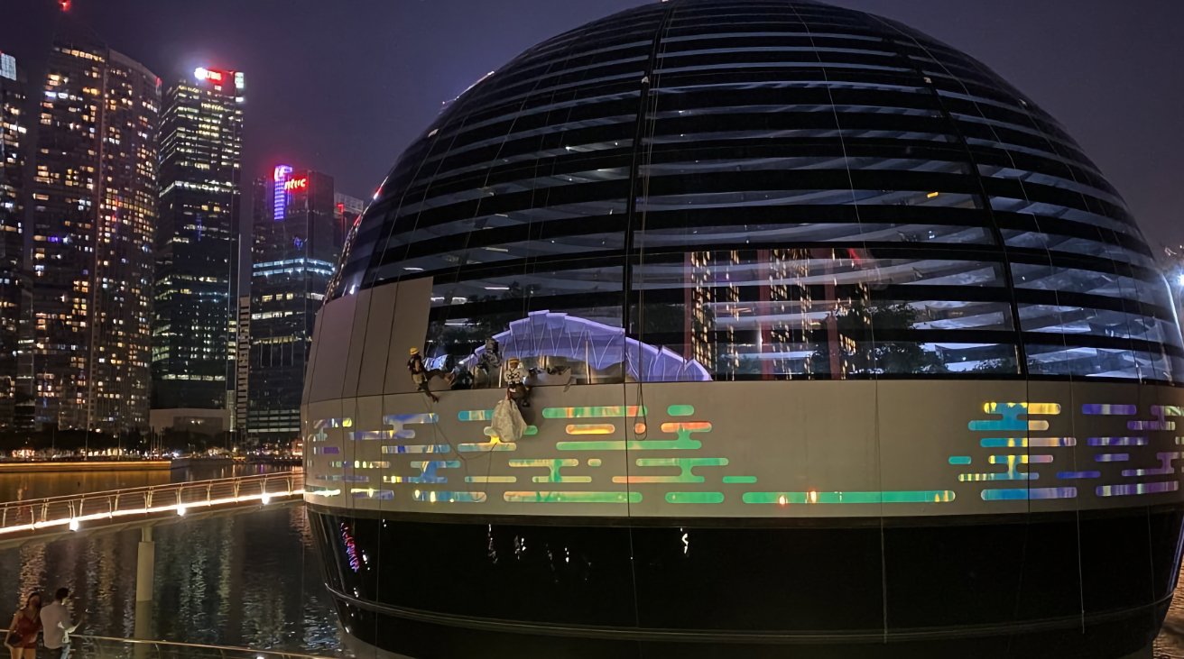 A spherical building lit with stripes and patterns against a night skyline, with people walking on a brightly lit waterfront promenade.