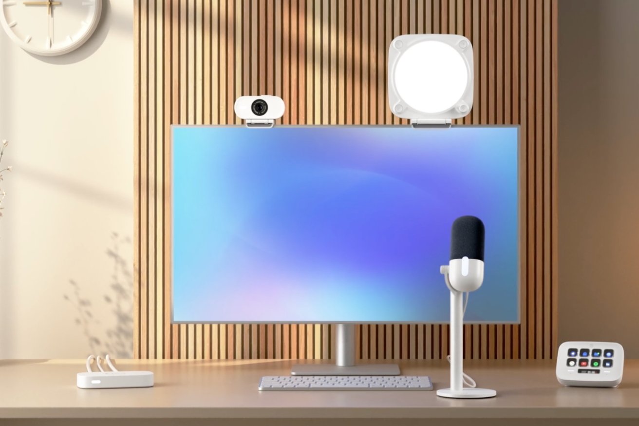 A modern desktop setup with a monitor, keyboard, webcam, microphone, smart speaker, and charging dock against a wooden slat background.