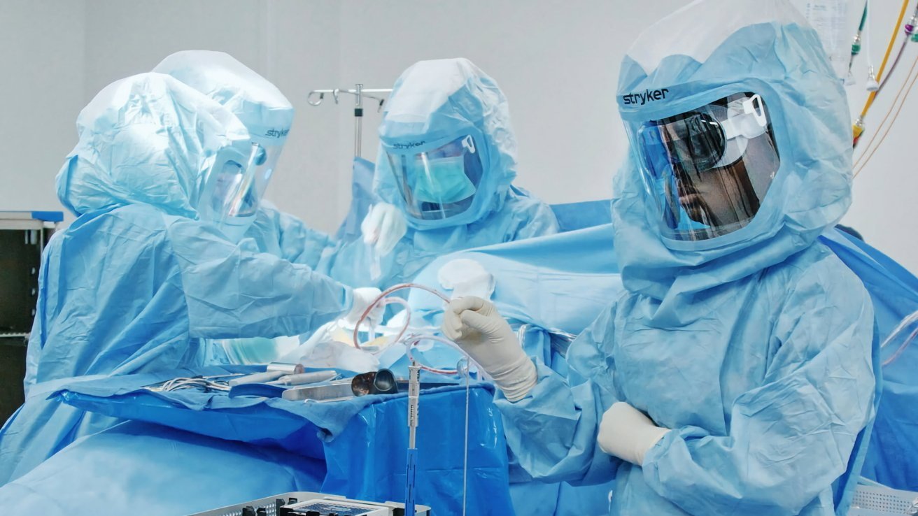 Three healthcare professionals in blue scrubs and protective gear operating surgical equipment in a medical setting.