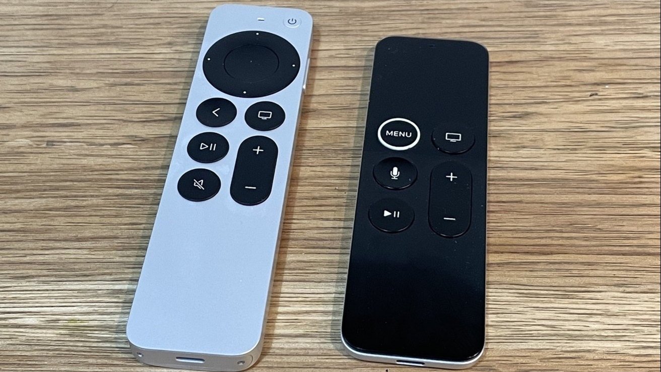 Two Siri Remote controls, one silver and one black, lying side by side on a wooden surface.