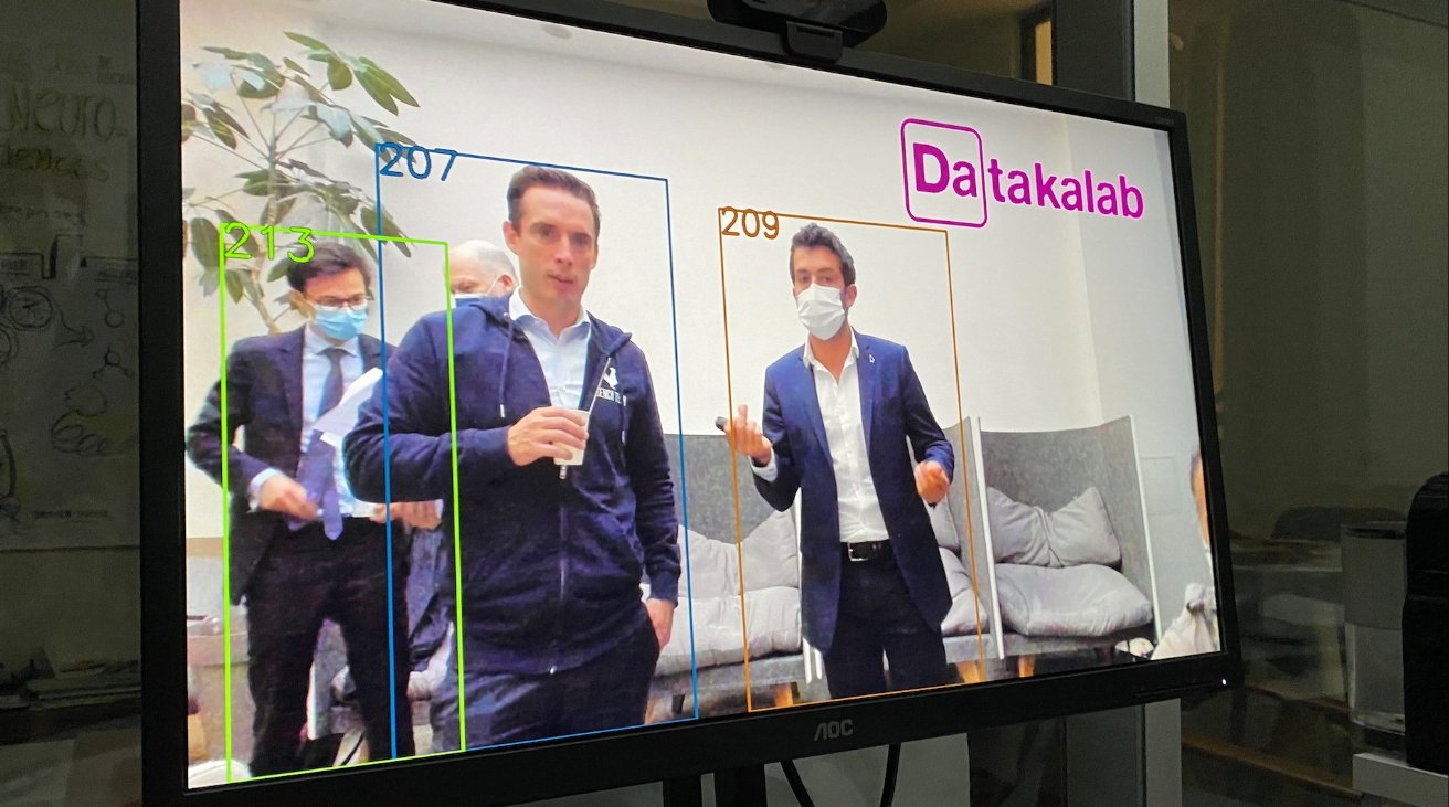 Monitor displaying three men in suits, one speaking, others listening, with face masks, in an office-like setting with annotations.