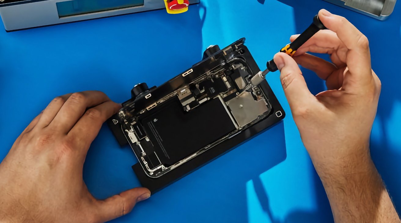Hands using a screwdriver to repair a disassembled electronic device on a blue surface.