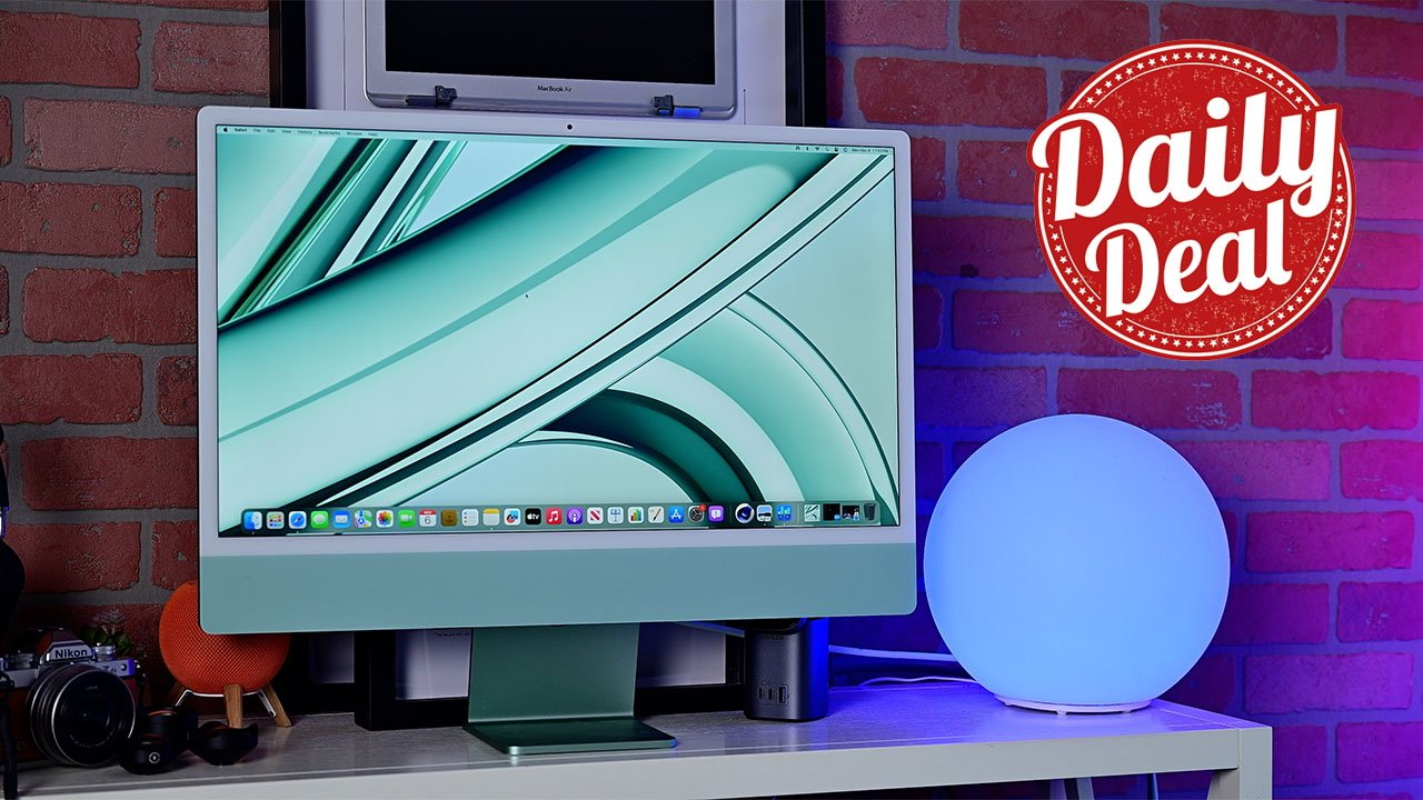 Apple's 24-inch iMac displaying a sleek interface, with a 'Daily Deal' sign, surrounded by various tech gadgets against a brick wall.