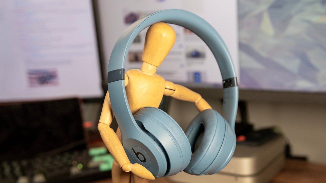 Wooden mannequin holding blue headphones against a blurred background with computer monitors.