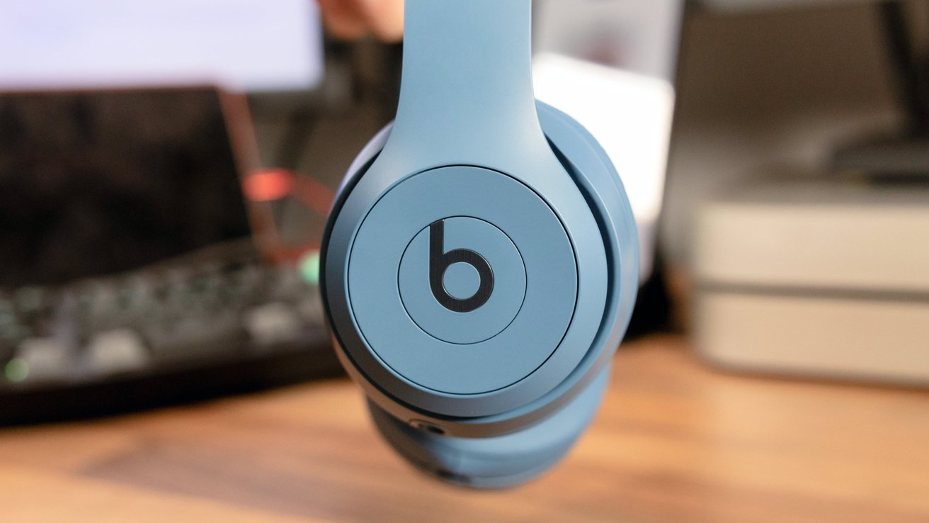 Close-up of a blue over-ear headphone with a circular logo on the side, blurry background with a desk and computer.