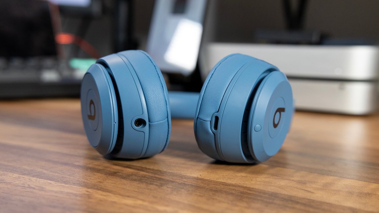 A pair of blue over-ear headphones resting on a wooden surface with electronic devices in the background.