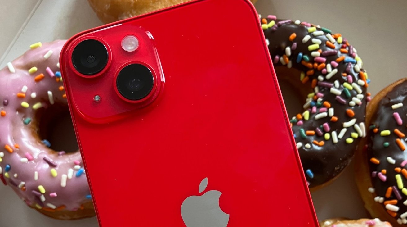 A red smartphone with dual cameras placed on a box of colorful sprinkle-topped donuts.