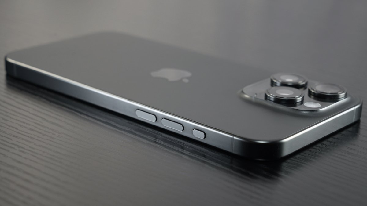 A close-up of an iPhone 15 Pro Max with a triple-camera setup on a textured surface.
