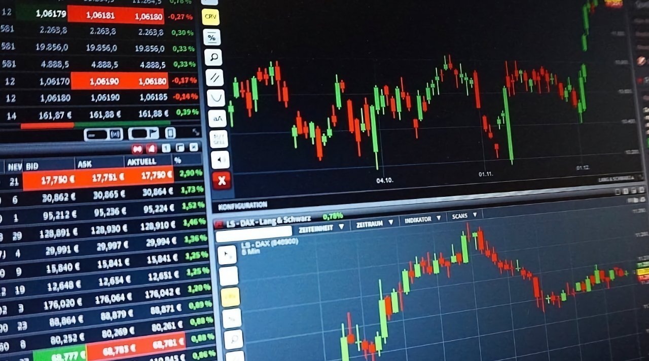 Multiple computer monitors displaying colorful financial trading charts and stock market data with red and green candlestick graphs.