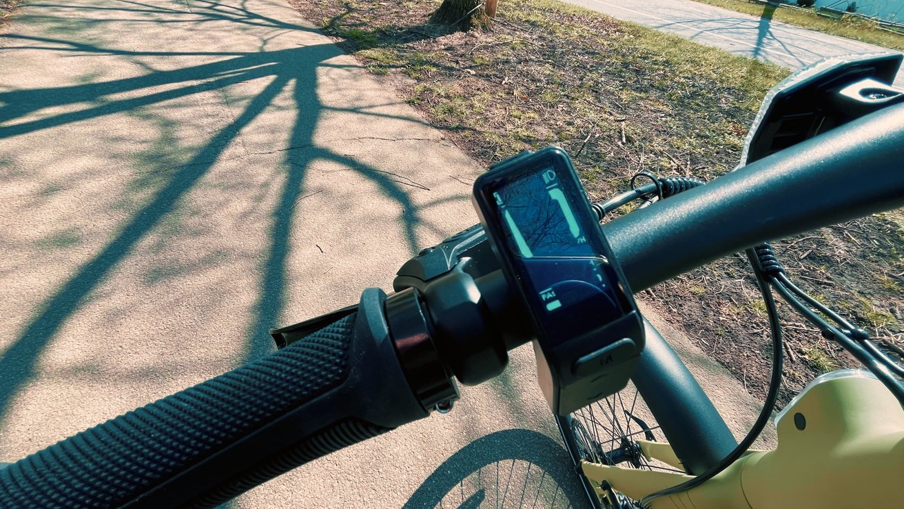 Handlebar view of a bicycle with a mounted display, casting a shadow on a sunny path.