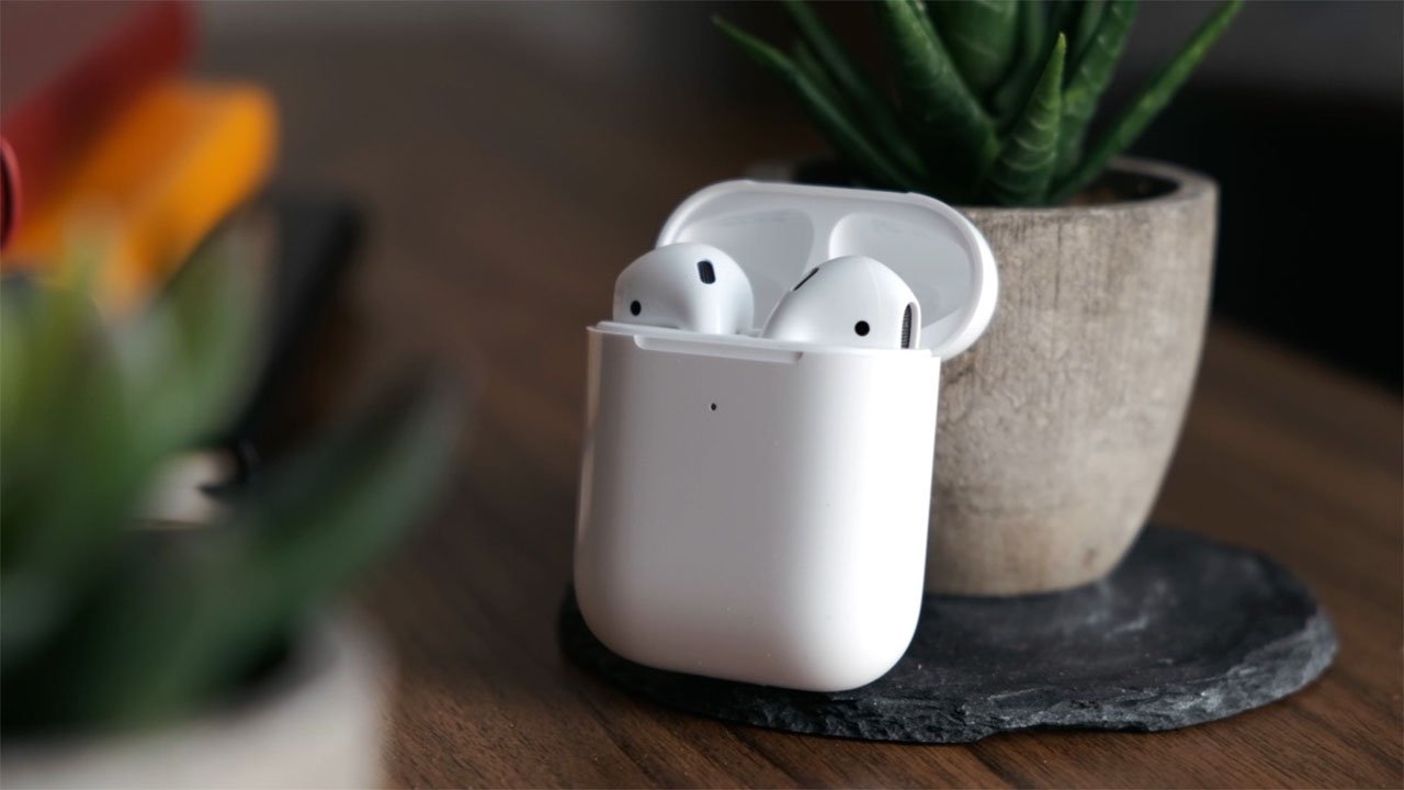 Apple AirPods 2 in its open charging case on a wooden table beside a potted plant.