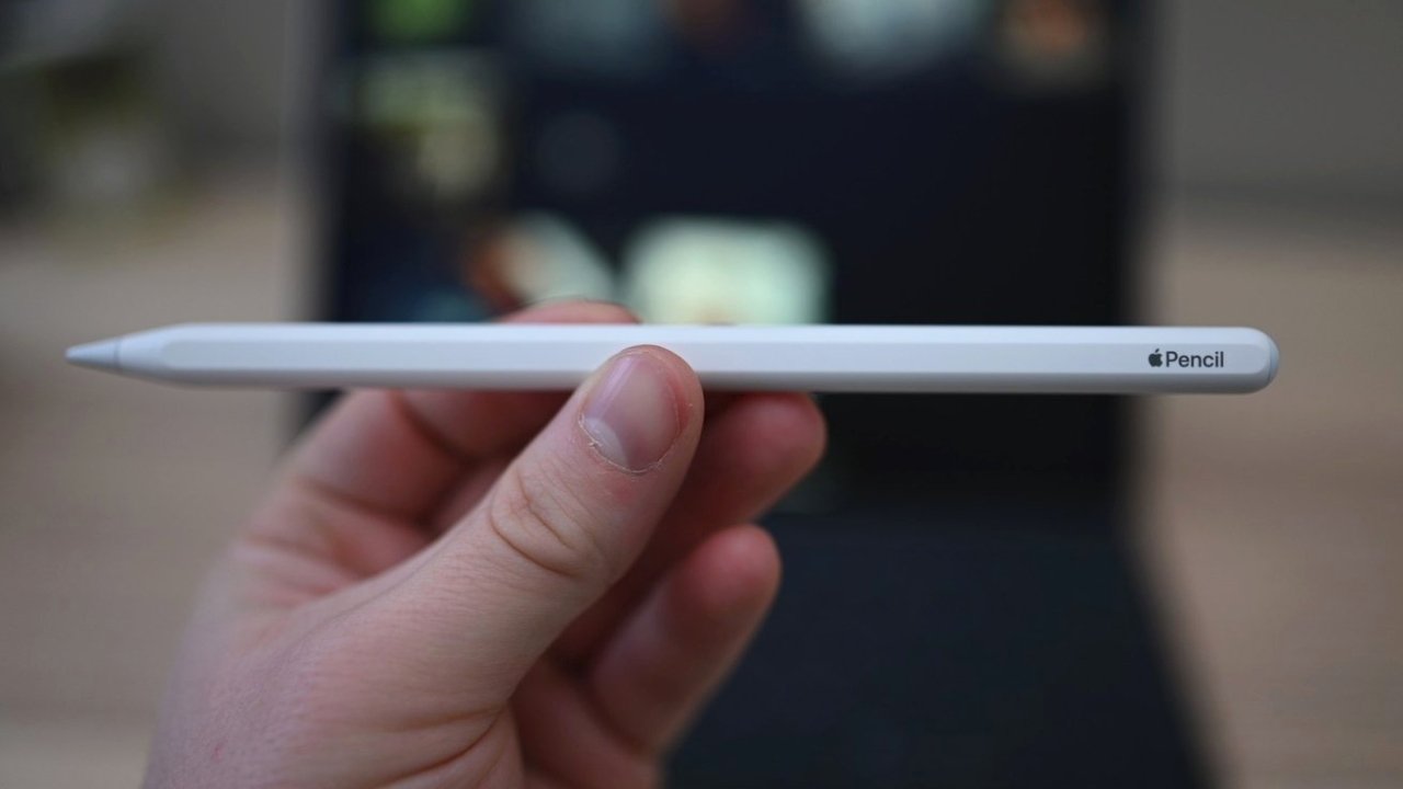 A close-up of a hand holding an Apple Pencil in focus, with a blurred background featuring a tablet.
