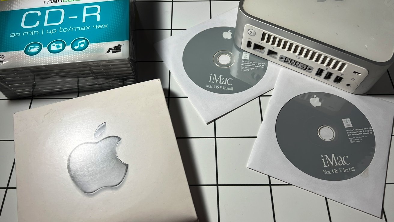 Apple products on a checkered surface, including iMac install CDs, CD-R pack, Mac OS box, and the back of a small desktop computer.