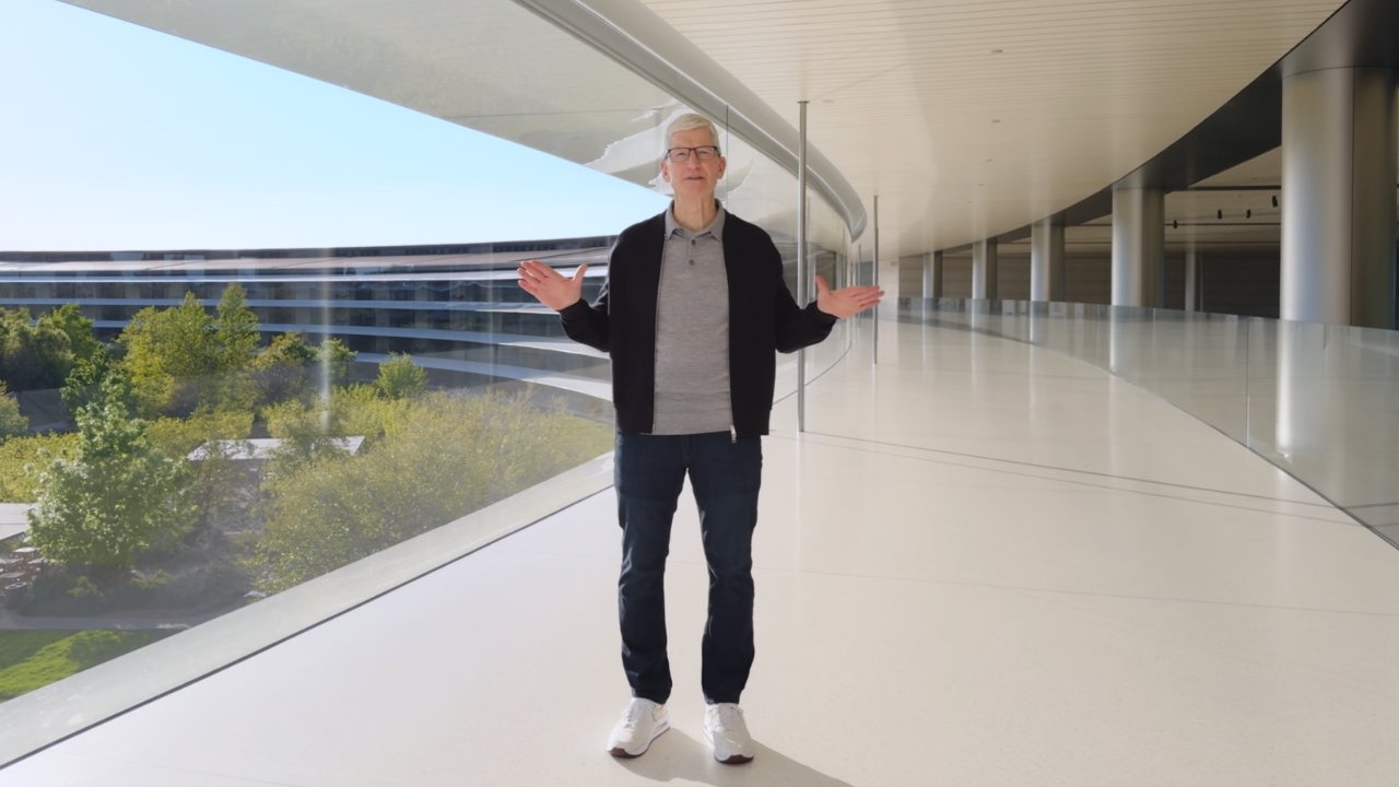 Man standing with arms outstretched in a modern hallway with glass walls overlooking green trees.
