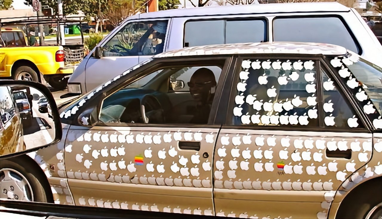A car covered in multiple apple-shaped stickers with a person visible through the window, parked beside a yellow vehicle.