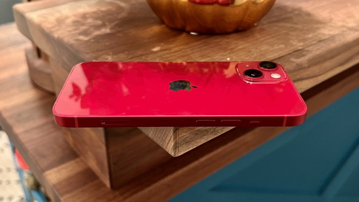 A red iPhone with a dual-camera system resting on a wooden surface, slightly overhanging the edge.