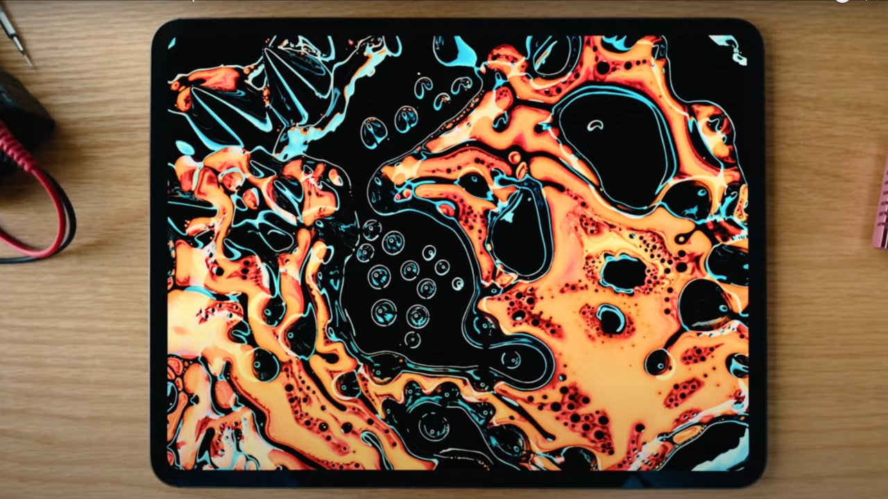 Apple iPad with a vibrant, abstract, orange and turquoise fluid art cover on a wooden desk next to a pink notebook.