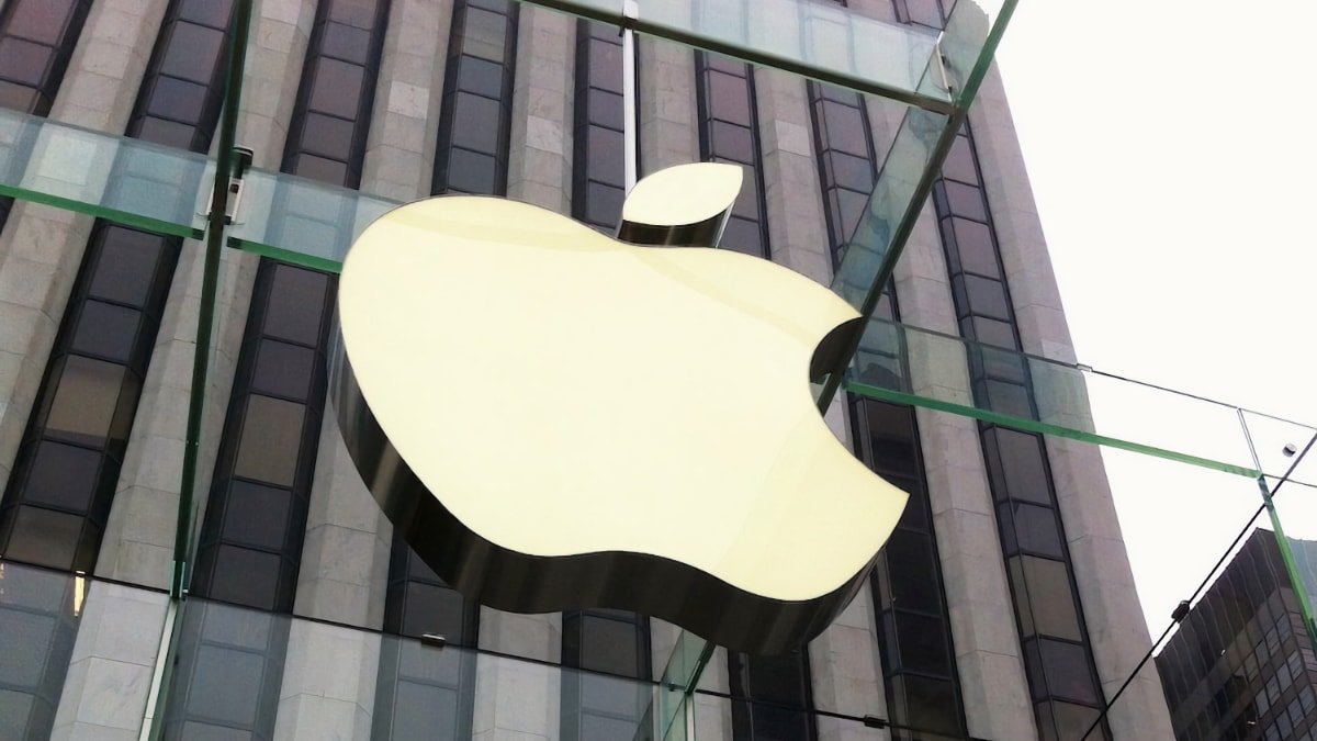 Large Apple logo suspended in front of a building with glass facade and overcast sky.