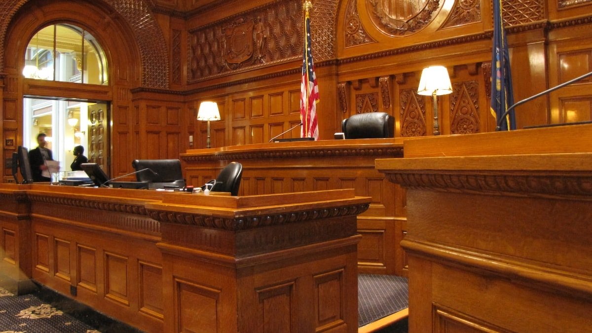 Ornate wooden courtroom with judge's bench, American flag, and two people in background.