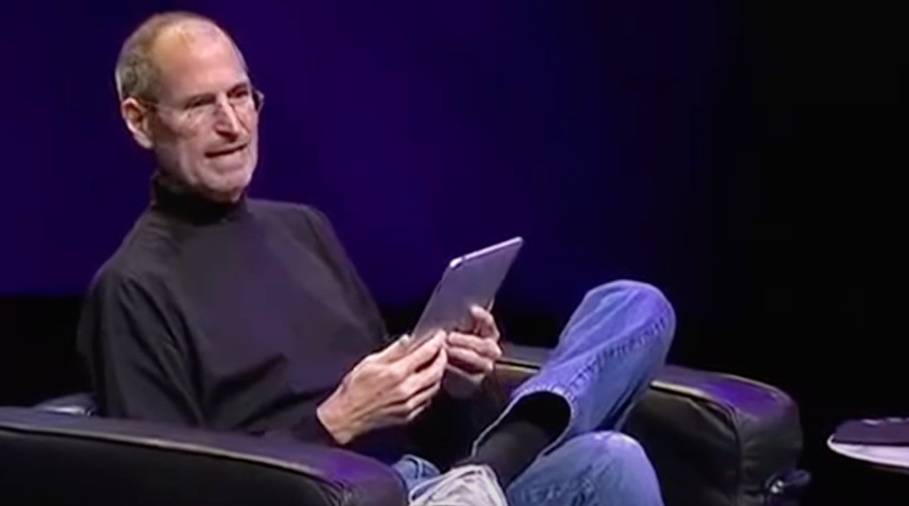 Man in black turtleneck and jeans sitting on a chair, holding and looking at a tablet on stage with dark background.