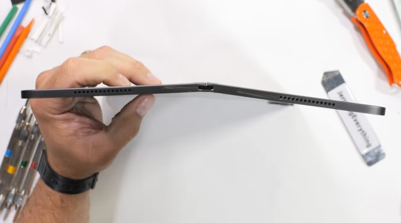 Side view of a bent iPad showing its ports, held by a person against a desk with disassembly tools.