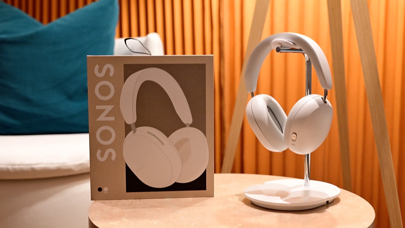 Sonos headphones displayed on a stand beside their packaging box on a table with a teal cushion and orange backdrop.