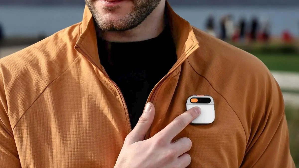 Man in a brown jacket pointing to a small white device clipped to his chest.
