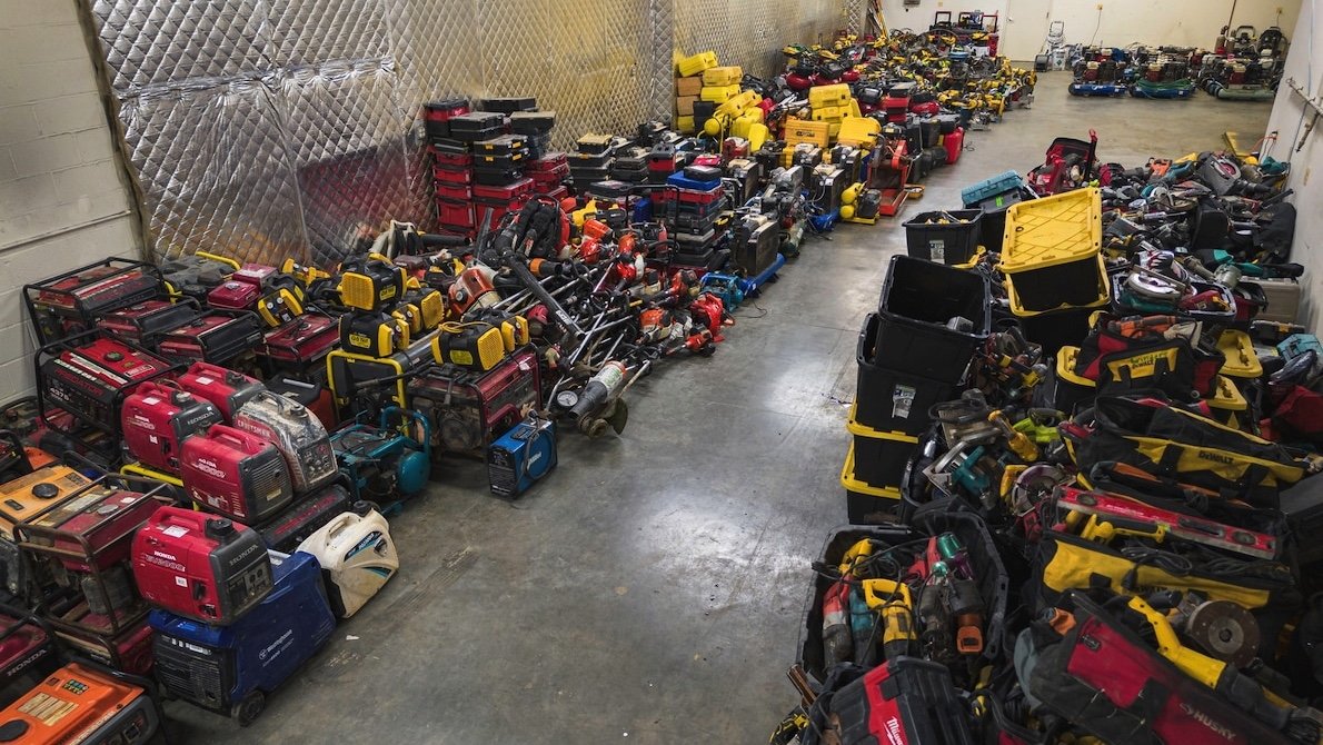 A cluttered warehouse with numerous power tools, generators, and equipment bags scattered across the floor and stacked on shelves.