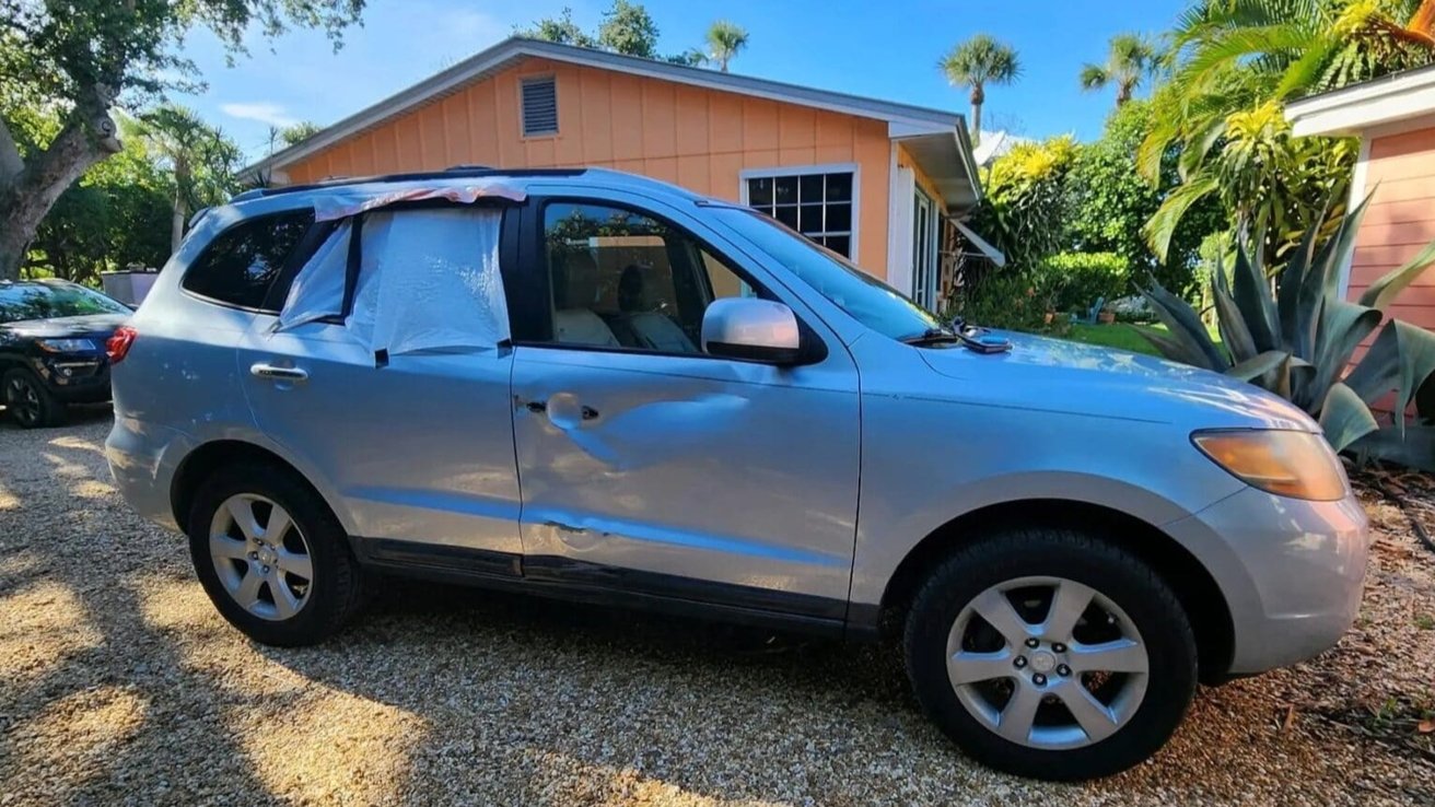 Silver SUV with plastic covering broken rear window and dents on the side, parked on gravel in front of an orange house surrounded by trees and plants.